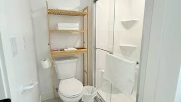 Une petite salle de bain comprenant des toilettes, une étagère en bois avec des serviettes et une cabine de douche en verre.