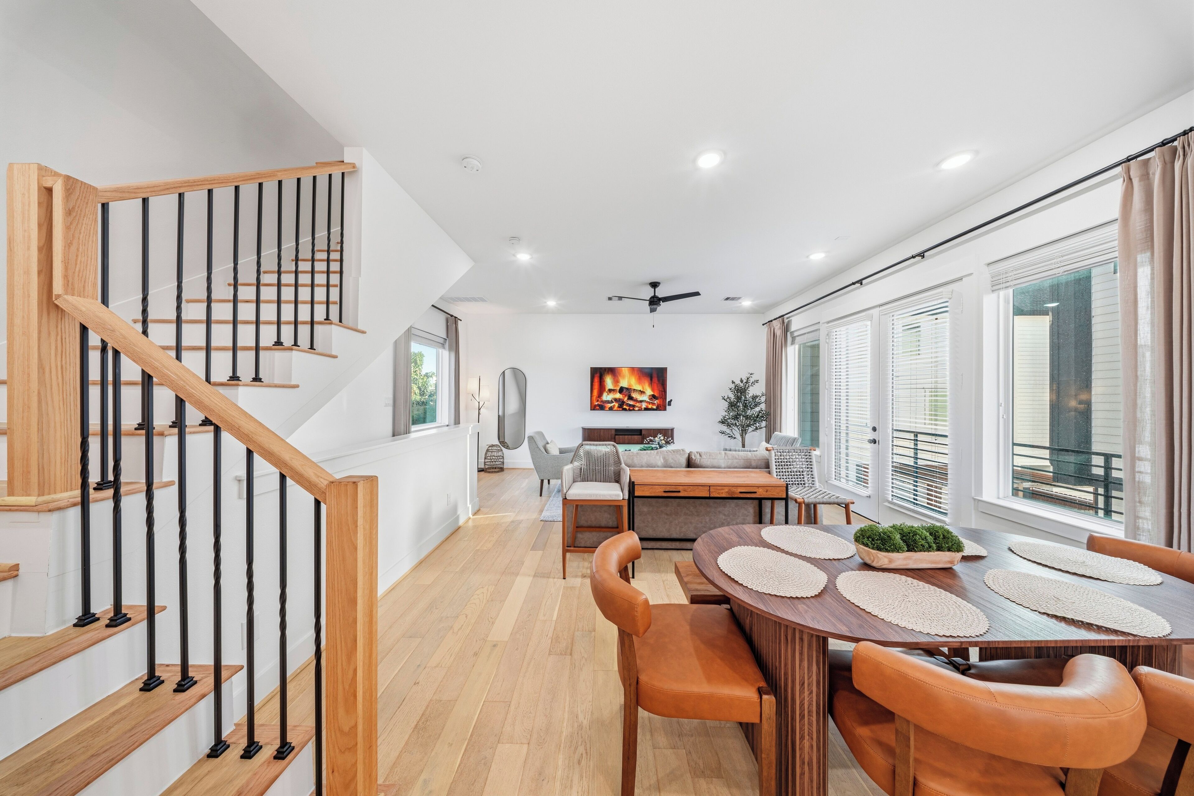 A modern interior with a wooden staircase, a dining area with a round table and leather chairs, and a large window with blinds.