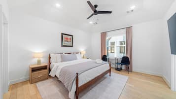 Un dormitorio con cama, mesitas de noche, un escritorio, una silla, ventilador de techo y ventana con cortinas.
