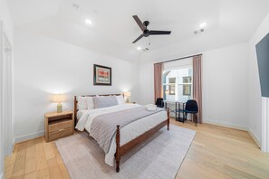 Ein Schlafzimmer mit einem Bett, Nachttischen, einem Schreibtisch, einem Sessel, einem Deckenventilator und einem Fenster mit Vorhängen.
