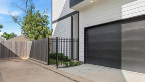 A modern two-car garage with a black door, a wooden fence, and a small garden area with plants. - Modern, luxe HTX Retreat| 8 Min to Toyota/Med Ctr (Houston)