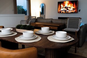 Ein Esstisch mit weißen Tellern und Schüsseln, ein Couchtisch aus Holz, eine Couch, ein Sessel und ein Fernseher, der ein Lagerfeuer zeigt.