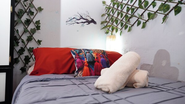 Un lit avec un oreiller rouge et un plaid coloré, un oreiller blanc et une couverture grise.