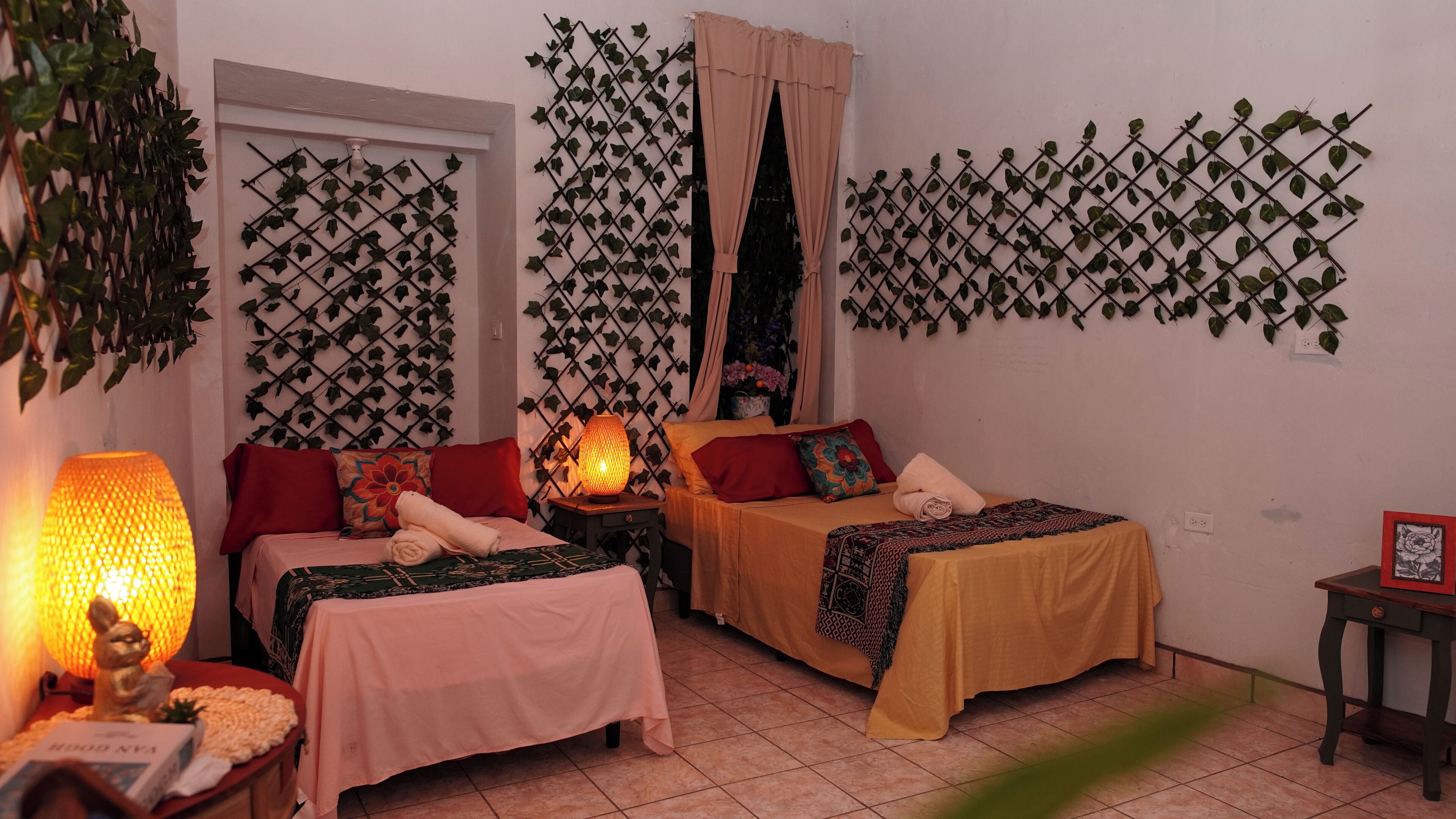 Una habitación con dos camas, una planta en la pared, una lámpara y una mesita con un libro.