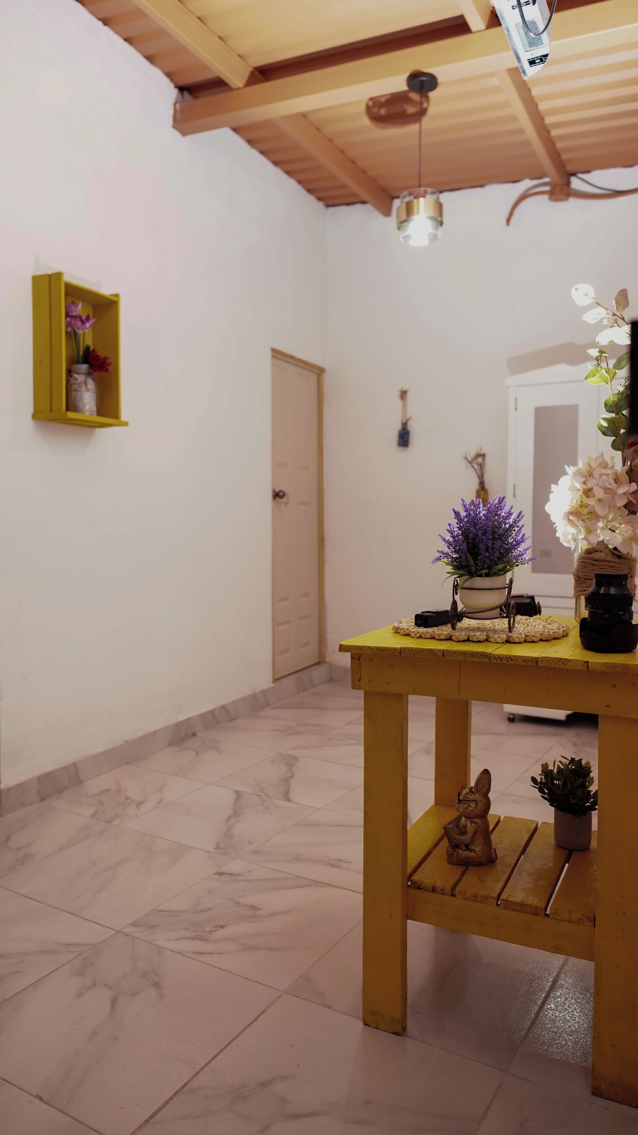 A room with a wooden table, a small shelf with flowers, a door, and a ceiling fan.