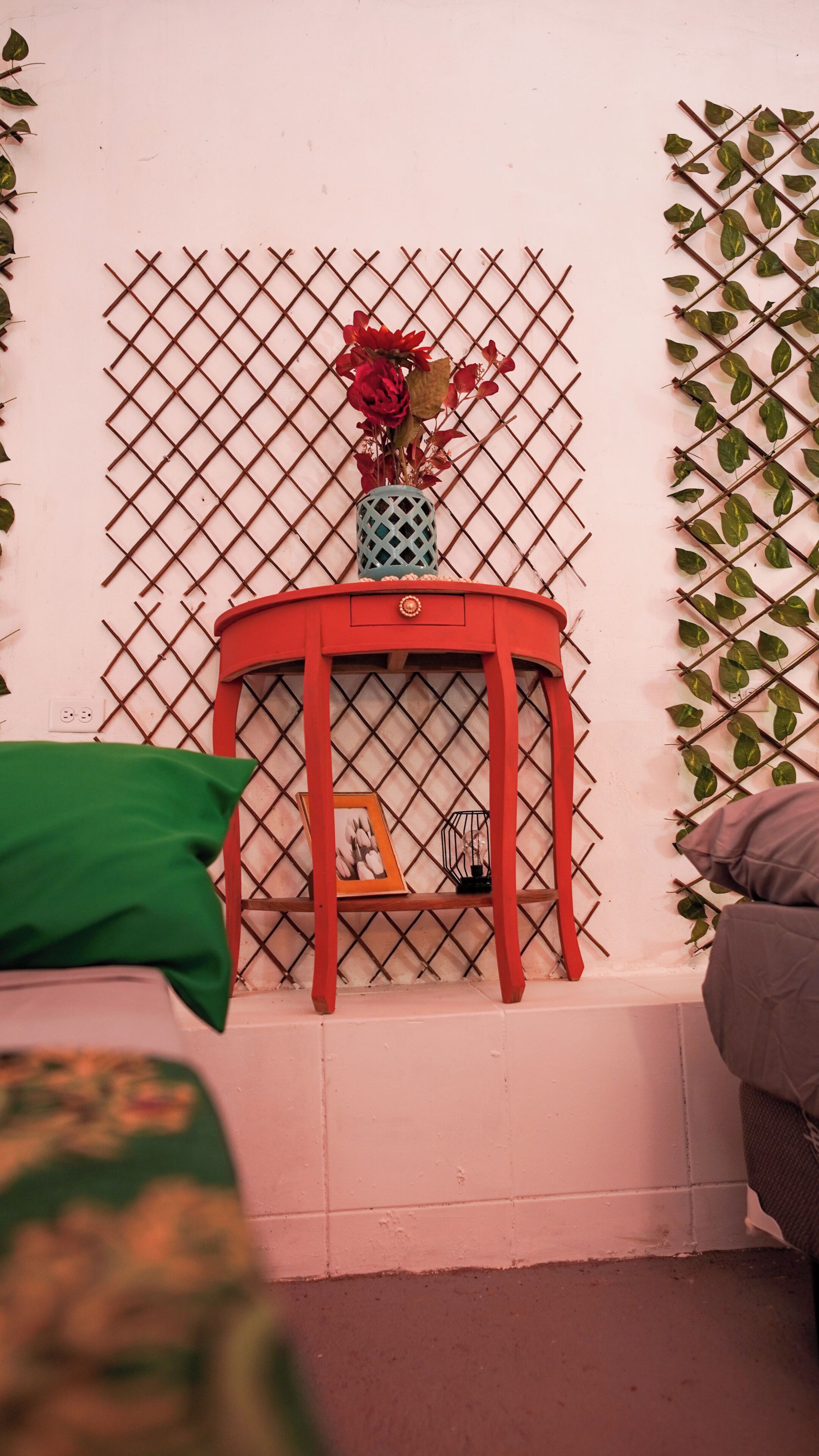 A room with a red stool against a wall with a grid pattern, a small shelf with a picture frame, and a green pillow.