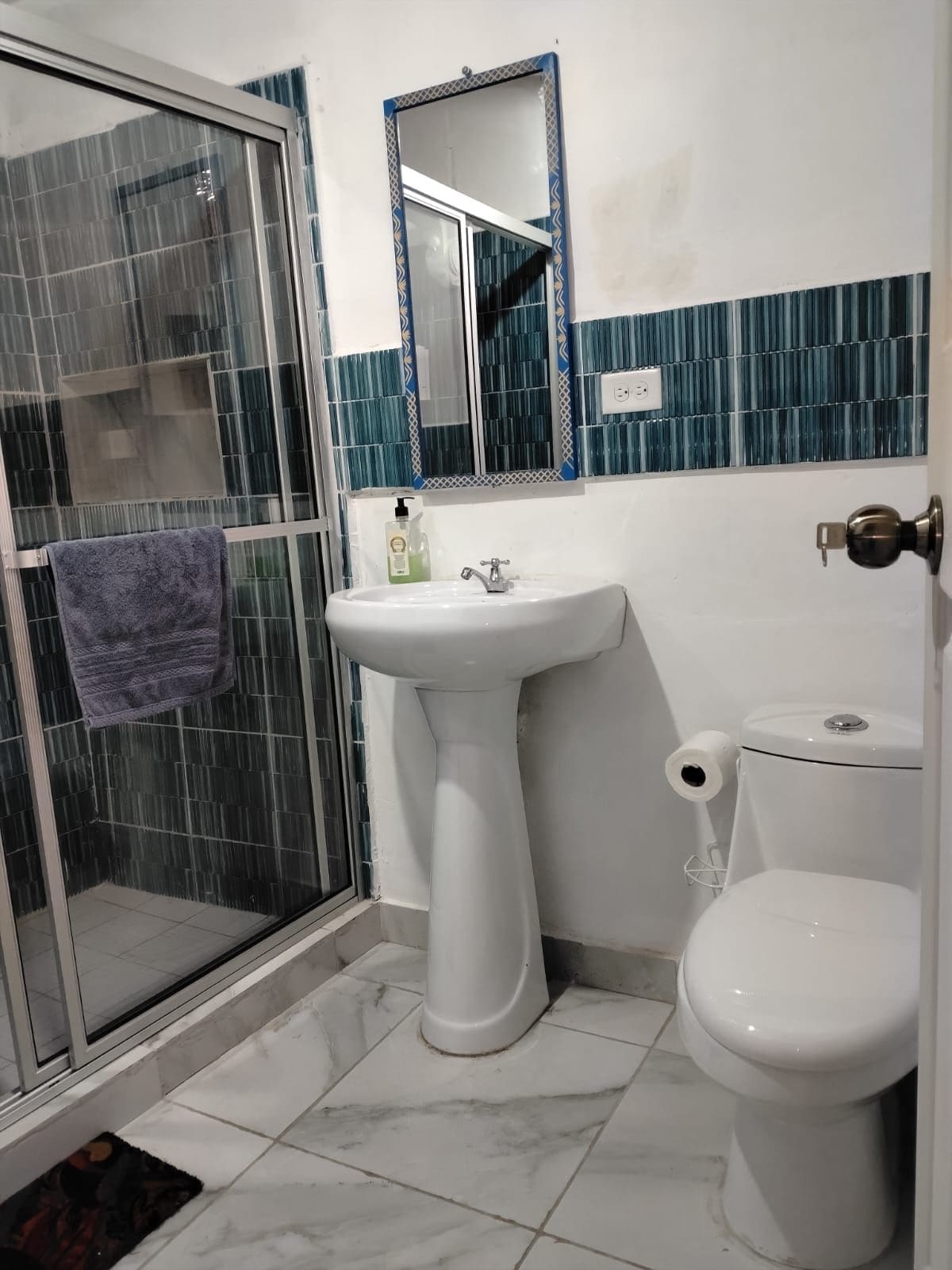 Un baño con cabina de ducha de vidrio, lavamanos sobre pedestal, inodoro y espejo sobre el lavamanos.