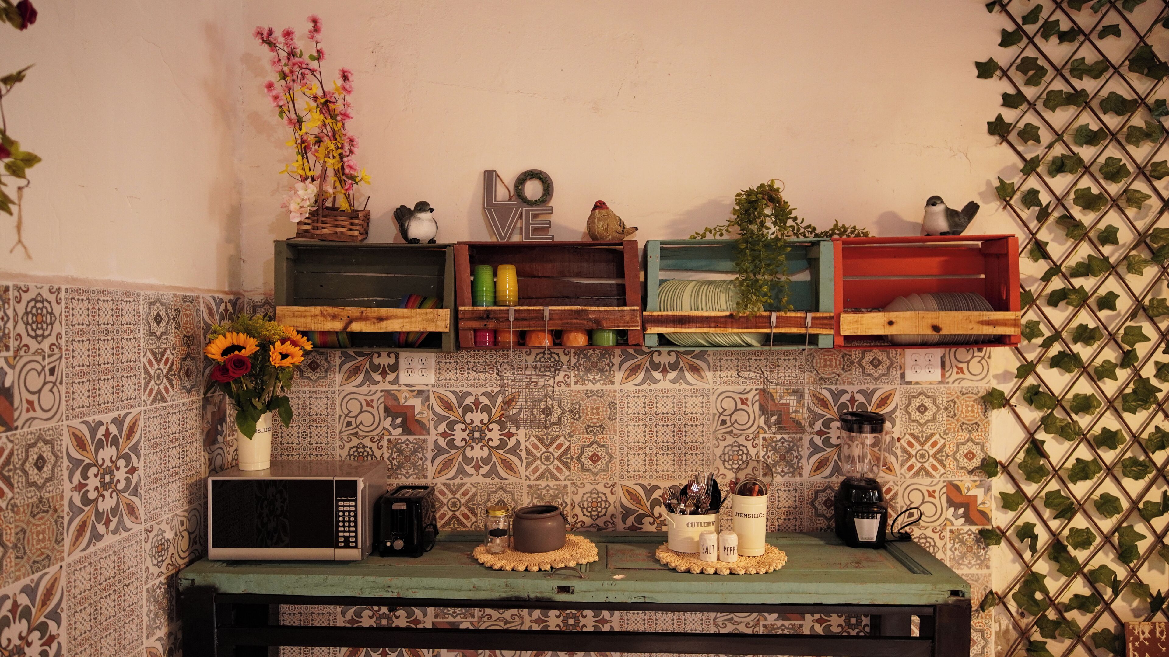 Una cocina con microondas, cafetera y estante con diversos objetos, incluyendo un jarrón con flores.