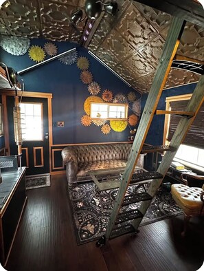 A room with a dark blue wall, a brown leather sofa, a wooden ladder, and a patterned rug. - Steampunk Tiny House w/ Loft Near Lake Travis (Austin)