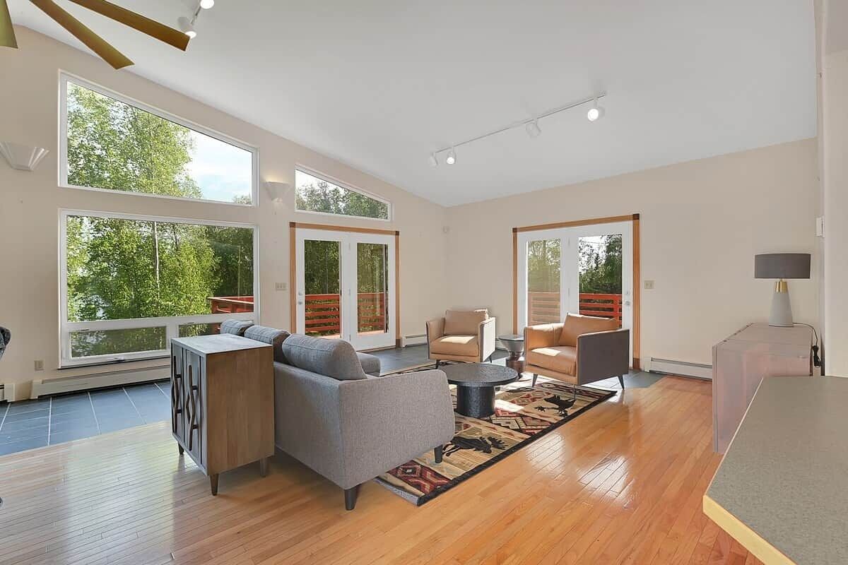 Un salon spacieux comprenant un canapé, des fauteuils, une table basse et un tapis. De grandes fenêtres laissent entrer la lumière naturelle et offrent une vue sur la verdure environnante.
