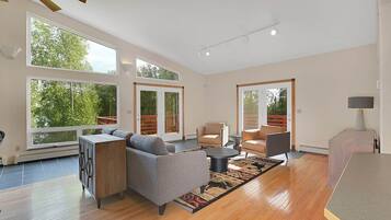 Un salon spacieux comprenant un canapé, des fauteuils, une table basse et un tapis. De grandes fenêtres laissent entrer la lumière naturelle et offrent une vue sur la verdure environnante.