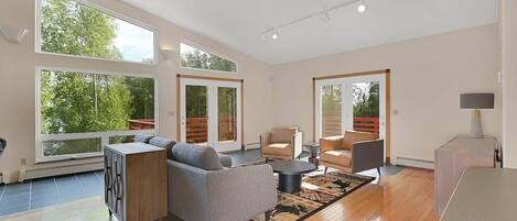 A spacious living room with a sofa, armchairs, a coffee table, and a rug. There are large windows providing natural light and views of greenery outside.