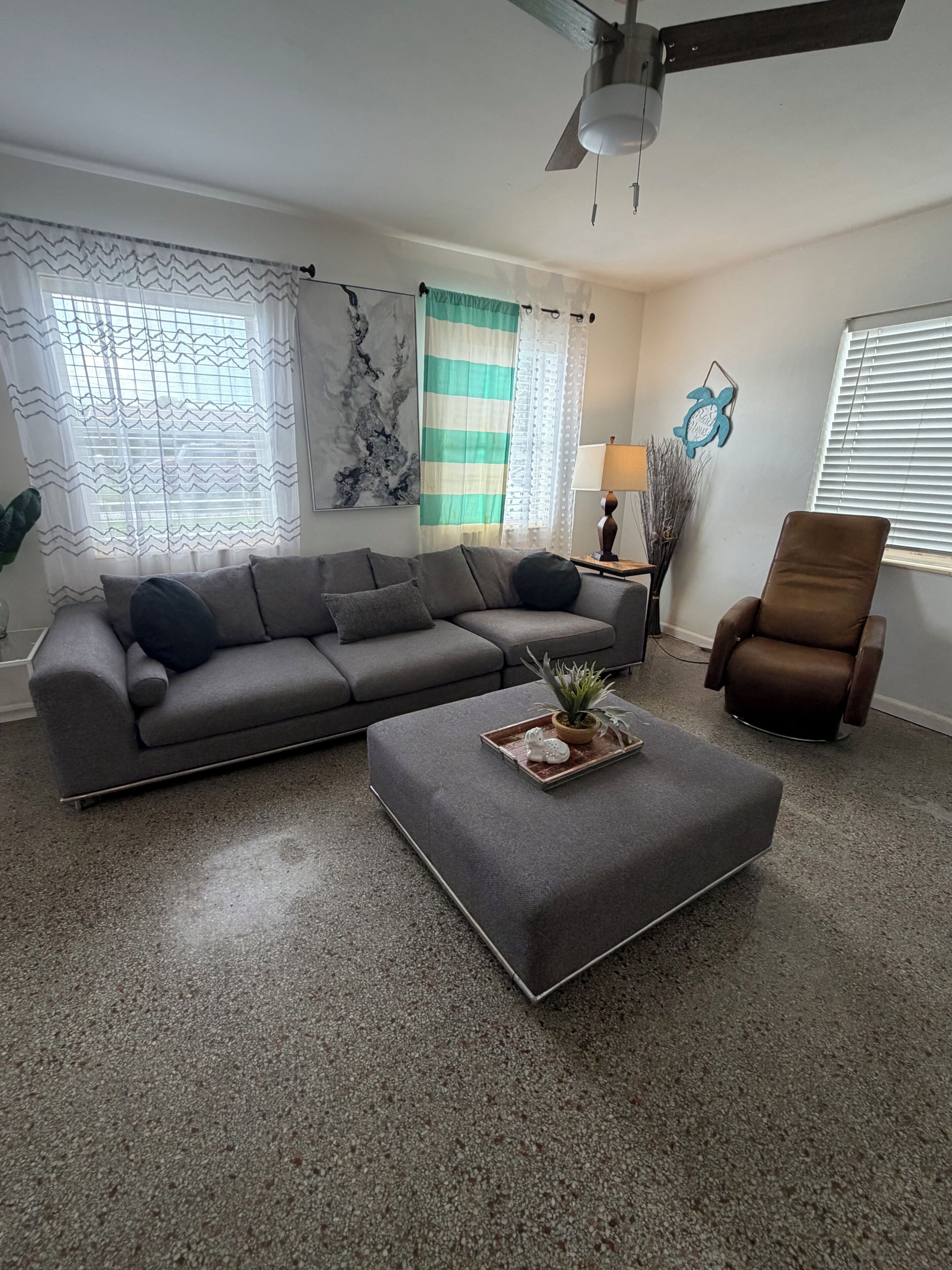 A living room with a grey sofa, a brown chair, a grey ottoman, and a ceiling fan.