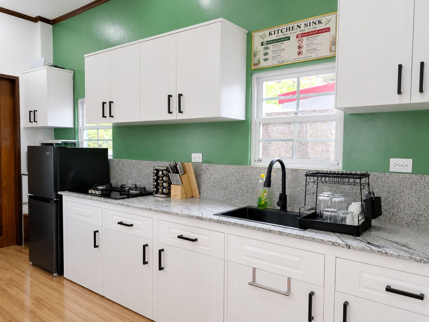 A kitchen with white cabinets, a black refrigerator, a sink with a black faucet, and a window with a view of greenery.