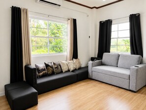 A modern living room with a grey sofa, black ottoman, and a black couch with patterned cushions. There are large windows with black curtains, and a wooden floor. - Modern home fast Starlink wi-Fi, Modern Home, free breakfast, near the beach (San Jose)