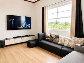 A modern living room with a flat-screen TV mounted on the wall, a black sofa with patterned cushions, a small ottoman, and a window with curtains.