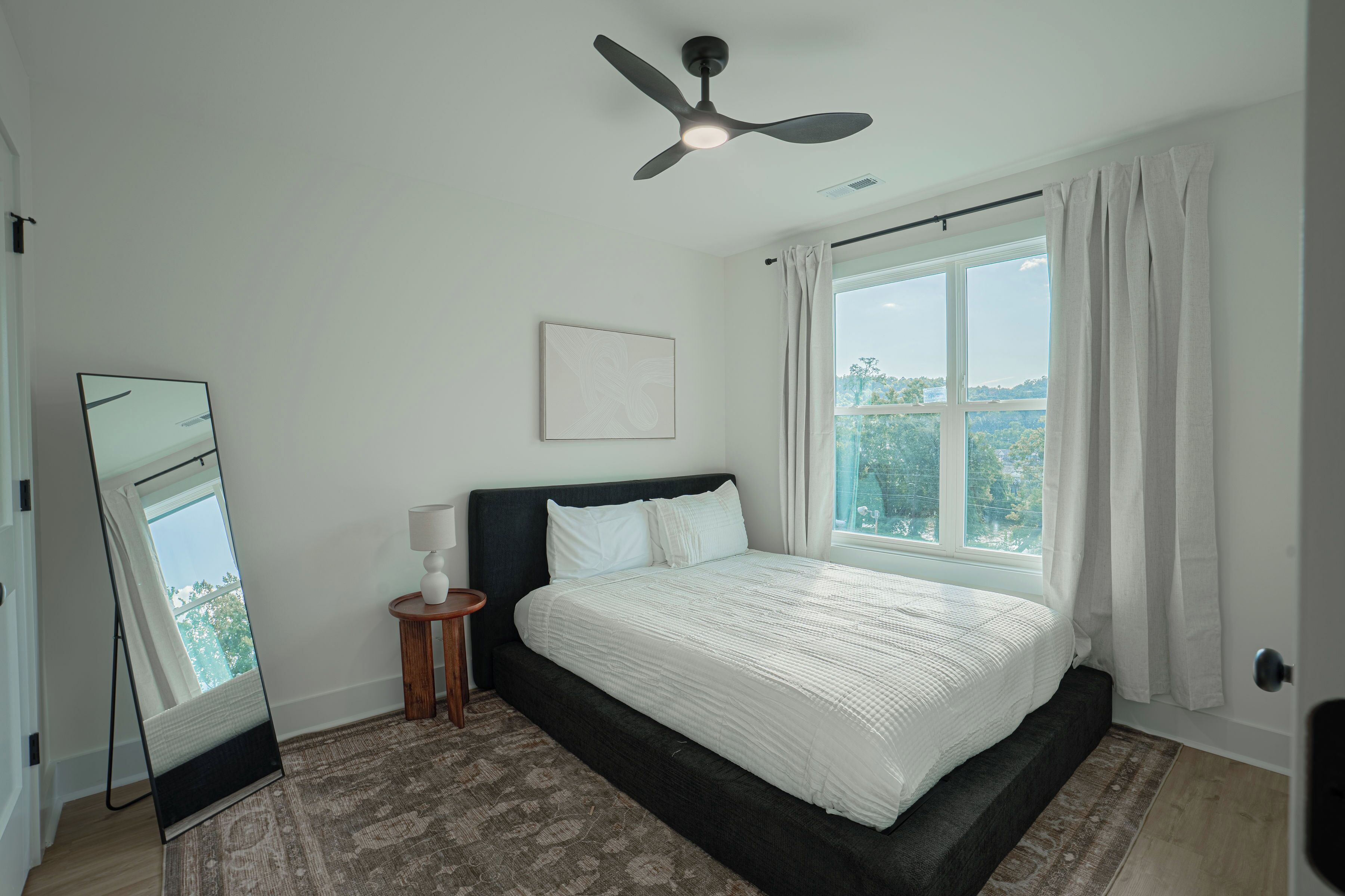 A bedroom with a bed, a ceiling fan, a window with curtains, a bedside lamp, and a framed picture on the wall.