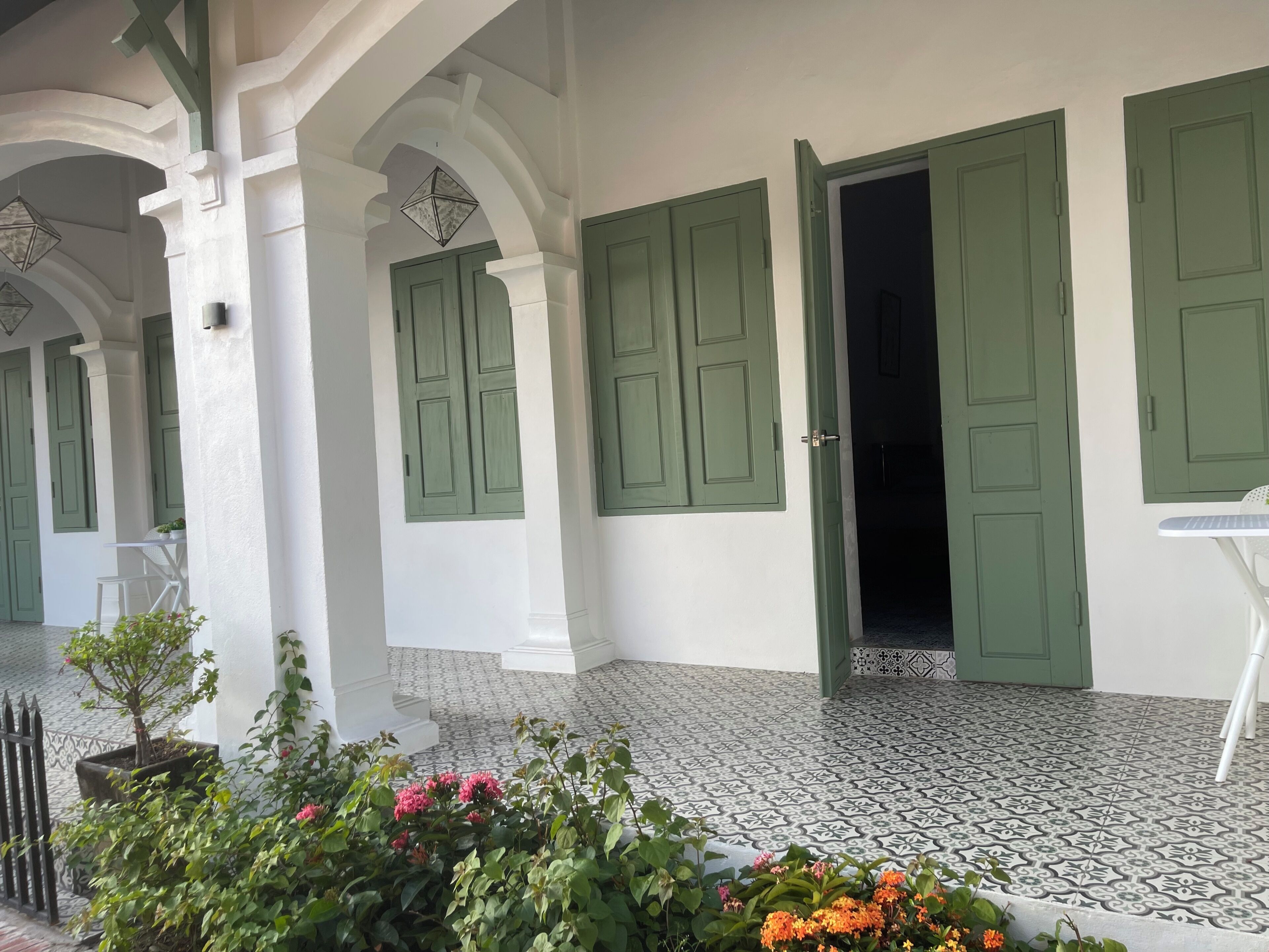 Un edificio con puertas verdes y paredes blancas, piso de baldosas y un pequeño jardín con flores.