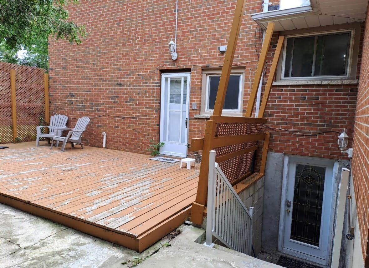 Een bakstenen gebouw met een houten terras, twee witte stoelen en een klein wit krukje.
