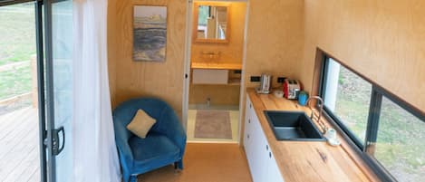 A compact kitchen with a large window, a blue chair, and a wooden floor.