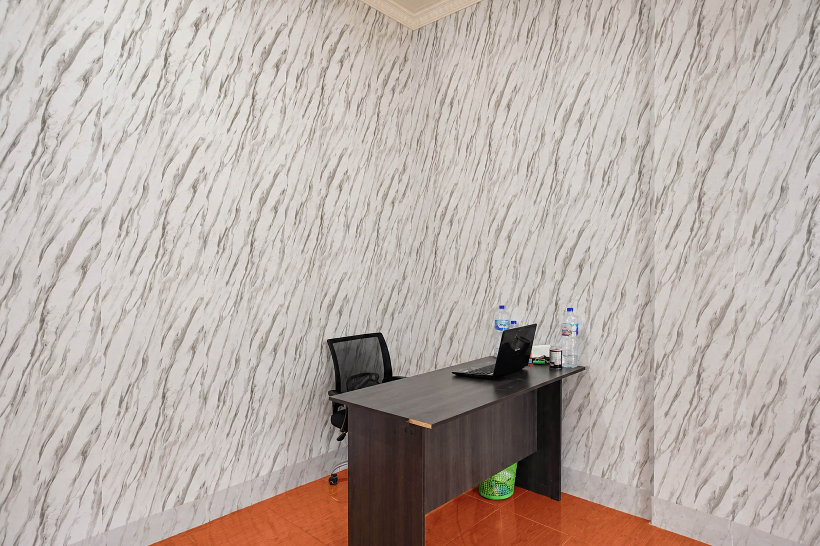 Una habitación con pared de diseño marmolizado, un escritorio de madera oscura, una silla de oficina negra y una botella de agua sobre el escritorio.