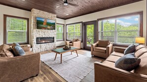 A spacious living room with a stone fireplace, leather sofas, and large windows. - Luxury Cabin w/Pool, Hot Tub, Game Room, Bar, Movie Room & Mountain Views (Sevierville)
