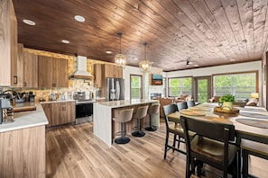 A modern kitchen with wooden cabinets, a central island, bar stools, and a dining area with a view of the outdoors. - The Ridgeline Reserve • Luxe Lodge w/ Private Indoor Pool • Top 5% (Sevierville)
