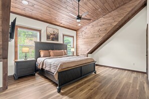 Ein Schlafzimmer mit schräg laufender Decke, einem Bett mit dunklem Kopfteil, zwei Nachttischen, einer Lampe und einem Deckenventilator.