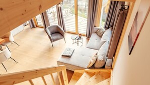 Un intérieur en bois avec un escalier menant à un balcon, un canapé gris avec des coussins, une petite table ronde avec une théière et une chaise blanche.