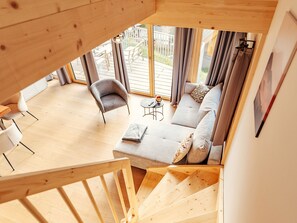 Ein Holzinnenraum mit einer Treppe, die zu einem Balkon führt, ein graues Sofa mit Kissen, ein kleiner runder Tisch mit einem Teekessel und ein weißer Stuhl.