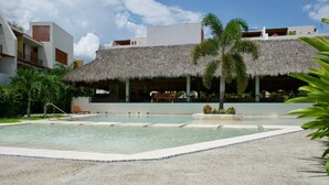 A pool area with a thatched-roof structure, palm trees, and a clear sky. - Modern Condo in Flamingos - Pool Gym Concierge (Bucerías)