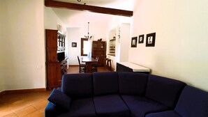 A living room with a dark blue sofa, a dining area with a wooden table and chairs, and a kitchen with cabinets and a chandelier. - Villa Carlebia - Private pool and garden. Beaches within walking distance, ideal for families (Sinnai)