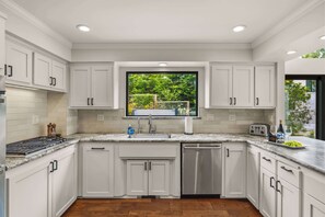 A modern kitchen with white cabinetry, a stainless steel dishwasher, and a window with a view of greenery. - Tide n Timber 4BR 2.5 BA (Pebble Beach)