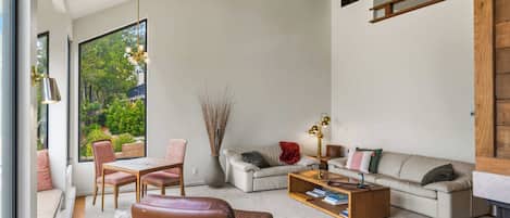 Una sala de estar moderna con sofá, mesa de centro, sillas, ventanas amplias y vista a zonas verdes.
