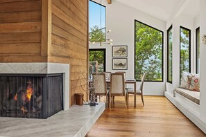 A modern interior with a fireplace, dining area, and large windows offering views of greenery. - Tide n Timber 4BR 2.5 BA (Pebble Beach)