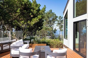 An outdoor seating area with white cushioned chairs and a wooden table, surrounded by trees and overlooking a body of water. - Tide n Timber 4BR 2.5 BA (Pebble Beach)