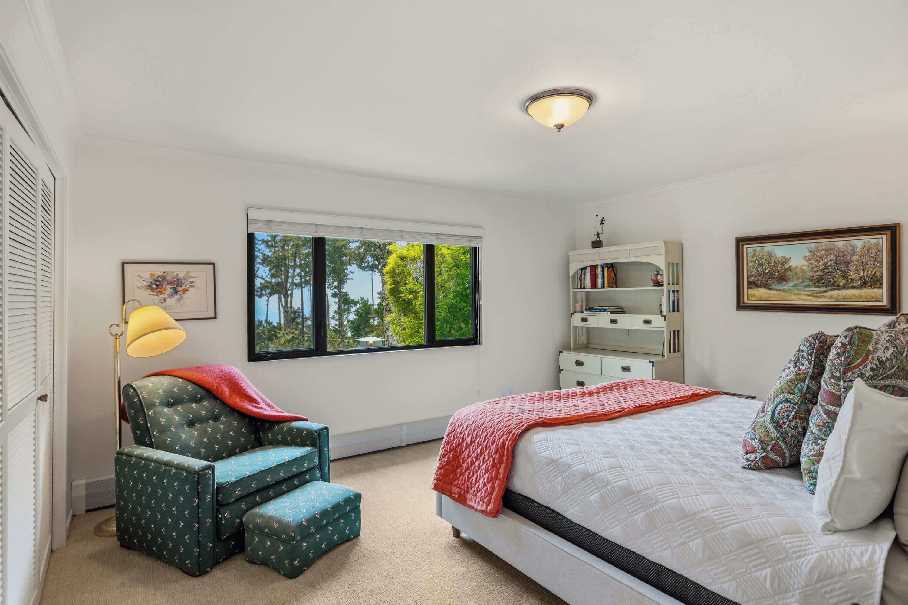 A bedroom with a bed, a chair, a desk, a window with a view of trees, and a ceiling light.