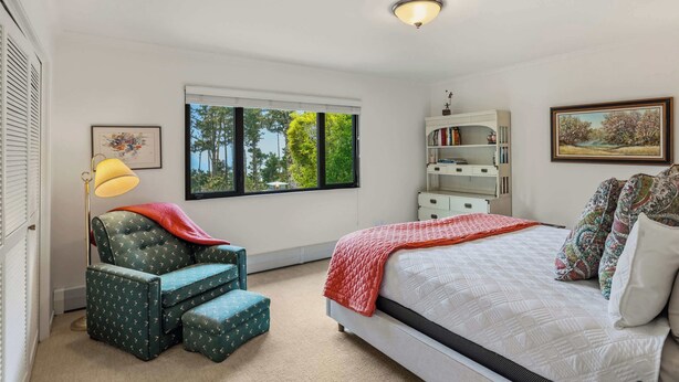 Ein Schlafzimmer mit einem Bett, einem Sessel, einem Schreibtisch, einem Fenster mit Blick auf Bäume und einer Deckenleuchte.