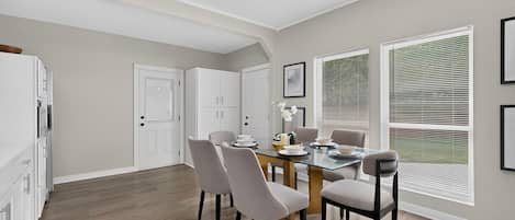 A modern kitchen with a dining area, wooden flooring, and large windows with blinds.