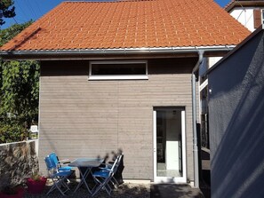 Ein modernes Gebäude mit einer grauen Fassade und einem roten Ziegeldach. Im Vorderbereich befindet sich ein kleines Fenster und eine Tür. Vor dem Gebäude steht ein Tisch mit Stühlen und ein Blumentopf.