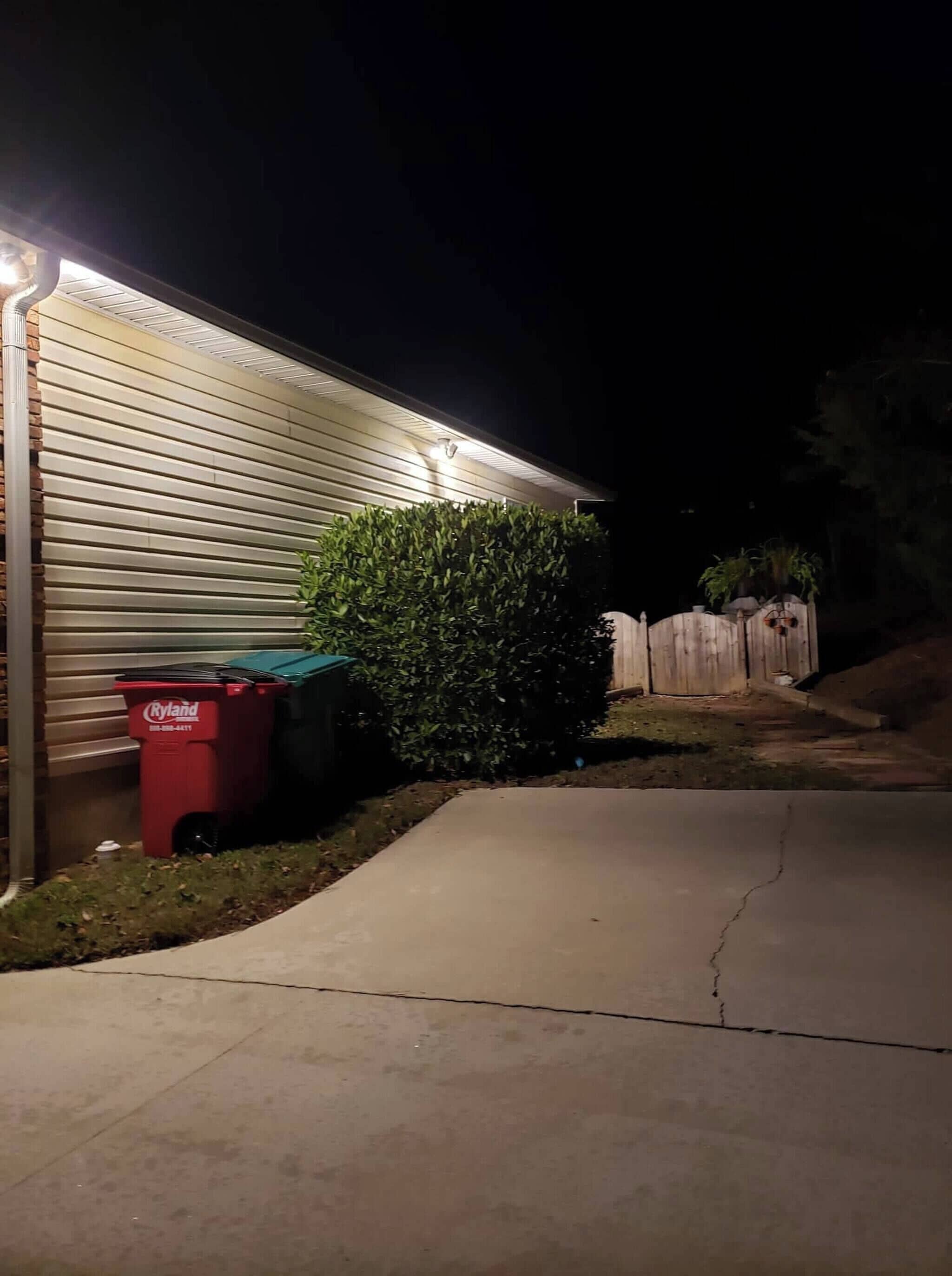 A residential driveway at night with a curb, a red trash bin, a green recycling bin, and a wooden fence.