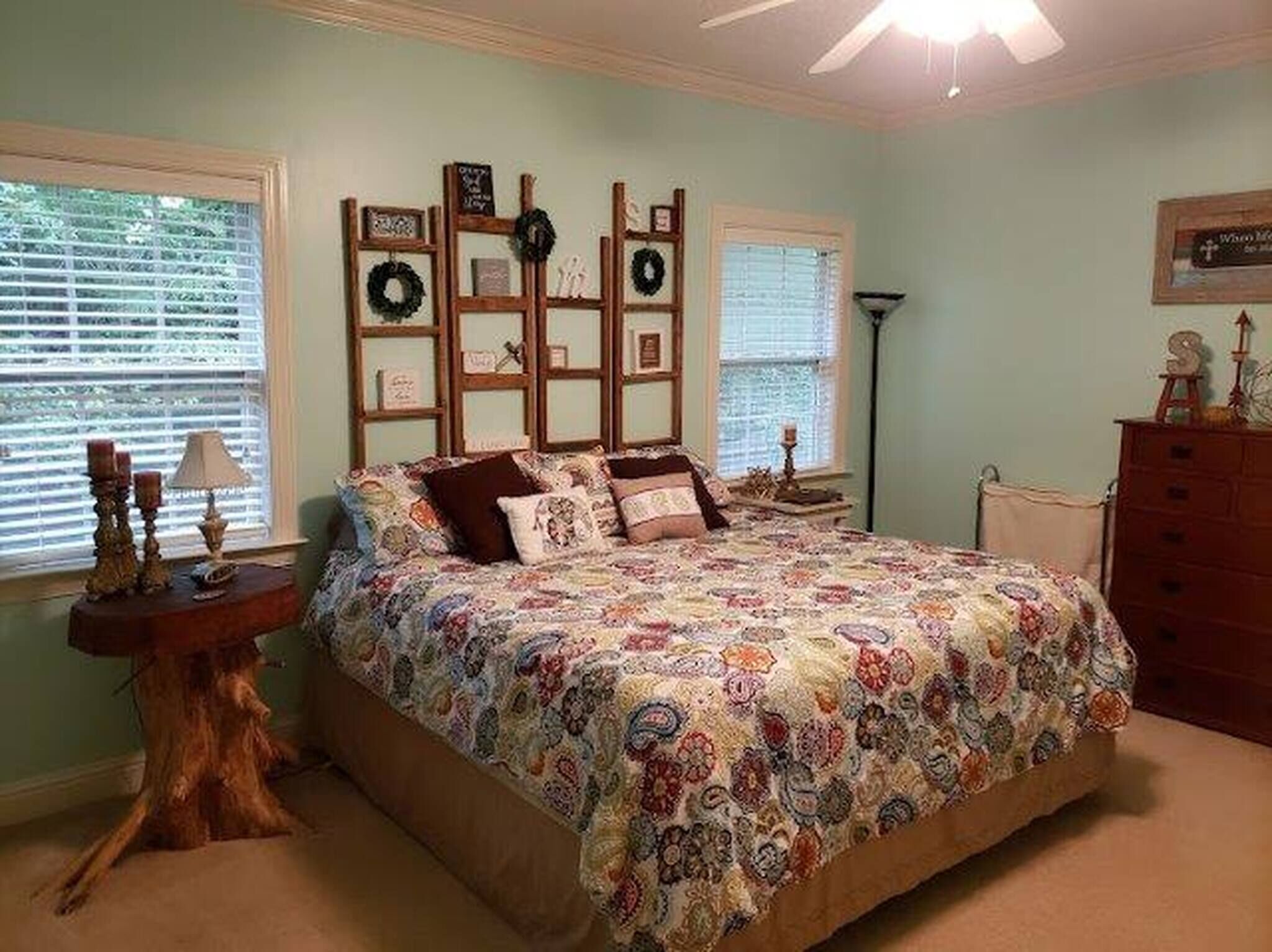 A bedroom with a large bed, a dresser, a nightstand, and a wall decorated with framed pictures and a wreath.