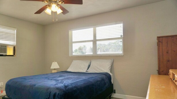 A bedroom with a bed, a ceiling fan, a window with blinds, and a wooden nightstand. - Plenty Space Bikers Sleep8 Beach (Ormond Beach)