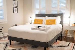 A neatly made bed with white bedding and two yellow pillows, a bedside table with a lamp, and a window with blinds. - The Mulberry at 121 Lincoln (Raleigh)