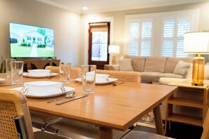 A dining area with a wooden table set for four, a television displaying a house, and a living room with a sofa and lamp. - The Mulberry at 121 Lincoln (Raleigh)