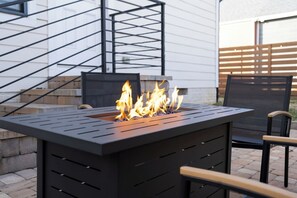 Ein schwarzes Aufeuerelement im Freien mit Flammen, ein schwarzer Metallstuhl und eine Holzbank.