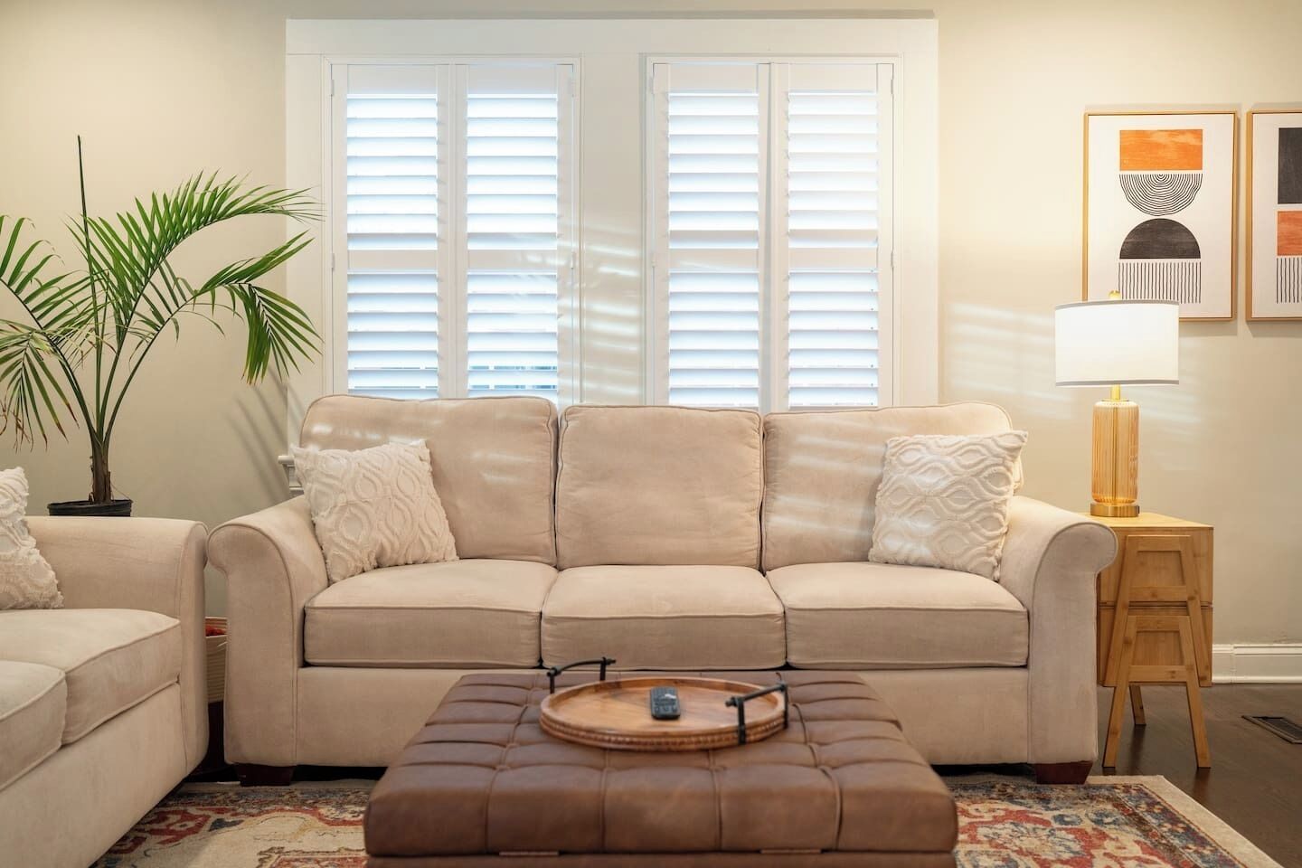 A living room with a beige sofa, a matching ottoman, a side table with a lamp, a potted plant, and a framed artwork on the wall.