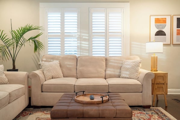 A living room with a beige sofa, a matching ottoman, a side table with a lamp, a potted plant, and a framed artwork on the wall. - The Mulberry at 121 Lincoln (Raleigh)