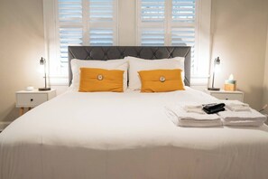 Ein ordentlich bezogenes Bett mit weißer Bettwäsche und zwei orangen Kissen, neben dem Nachttische mit Lampen und einer Taschentuchbox stehen.