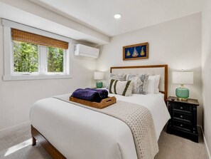 A bedroom with a bed, bedside tables, a window with blinds, and an air conditioning unit.